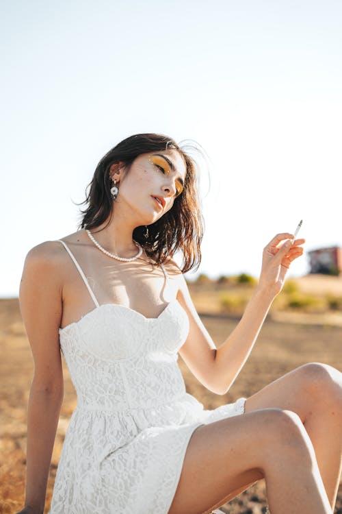 Young Woman in a White Spaghetti Strap Mini Dress Sitting in a Field and Smoking a Cigarette