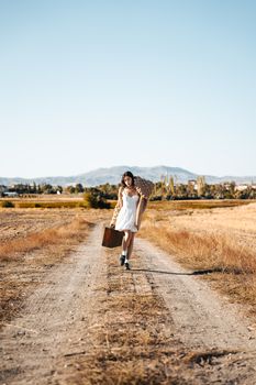 A young woman in a white dress and beige sweater carries a suitcase on a dirt road in the countryside.