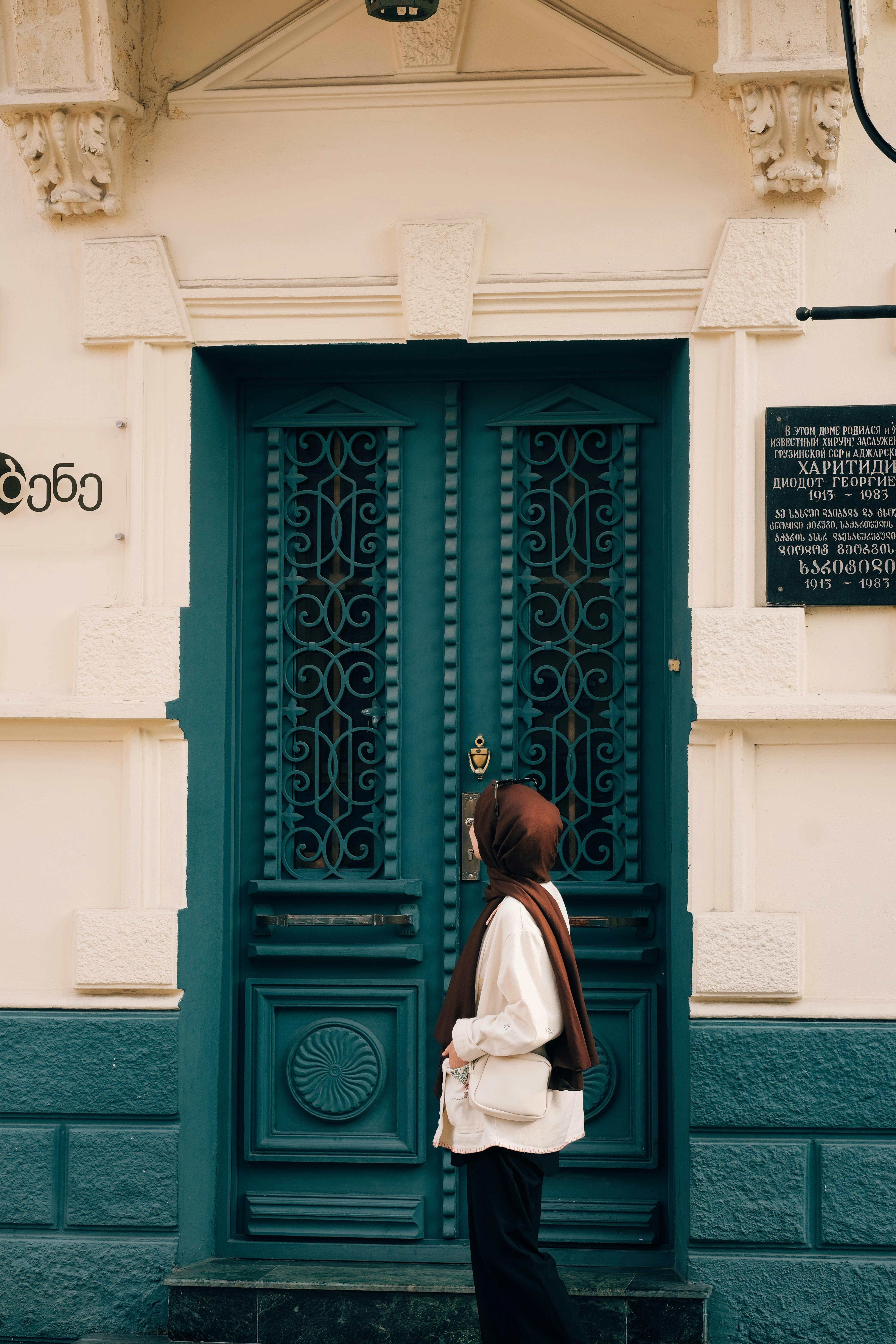 A woman in a headscarf stands by an ornate teal door on a building facade.