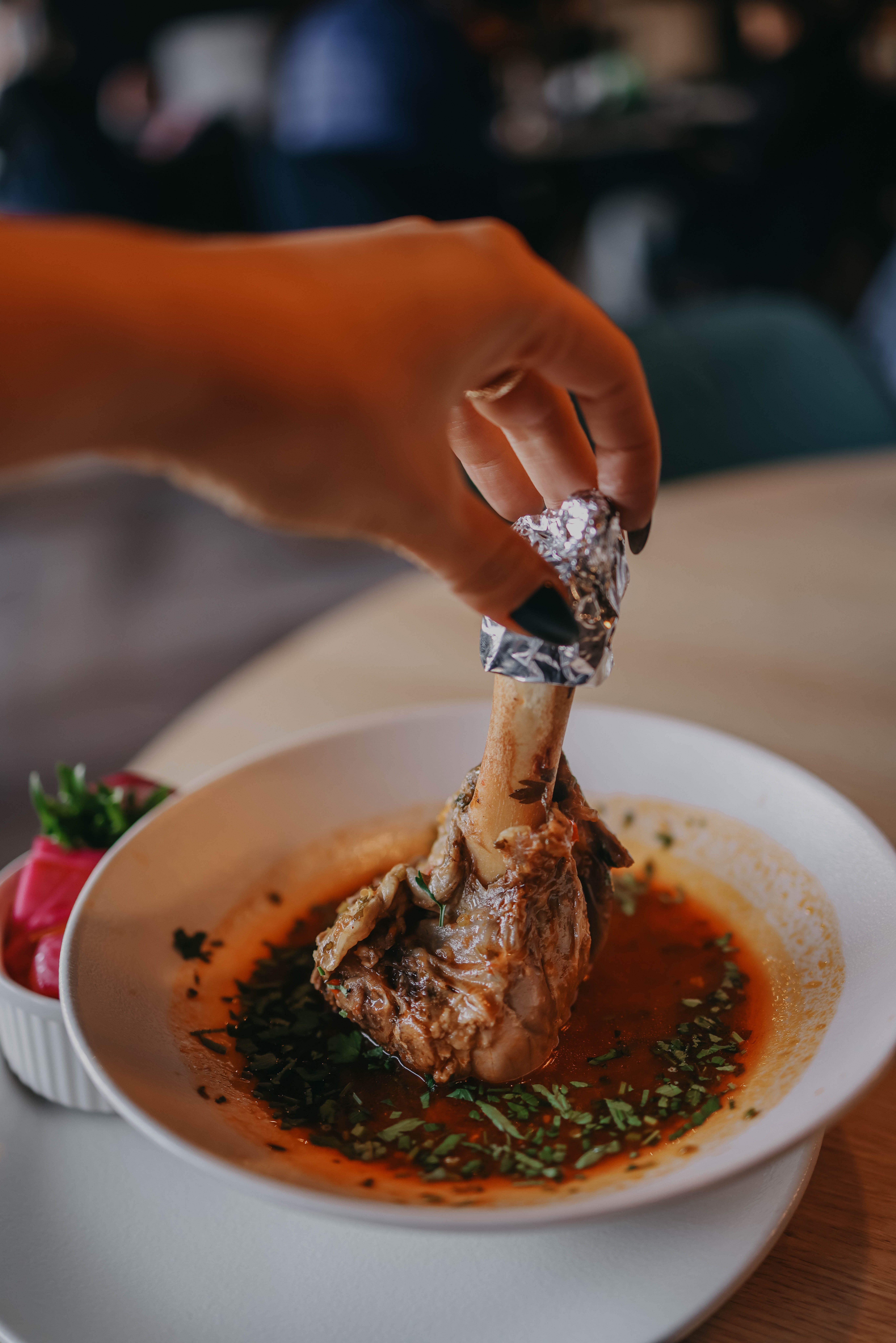 Hand holding a lamb shank in a flavorful broth, garnished with herbs.