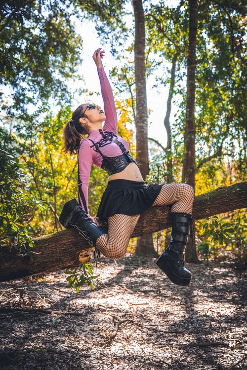 Free Woman in goth style attire striking a pose on a fallen tree trunk in a sunny forest setting. Stock Photo