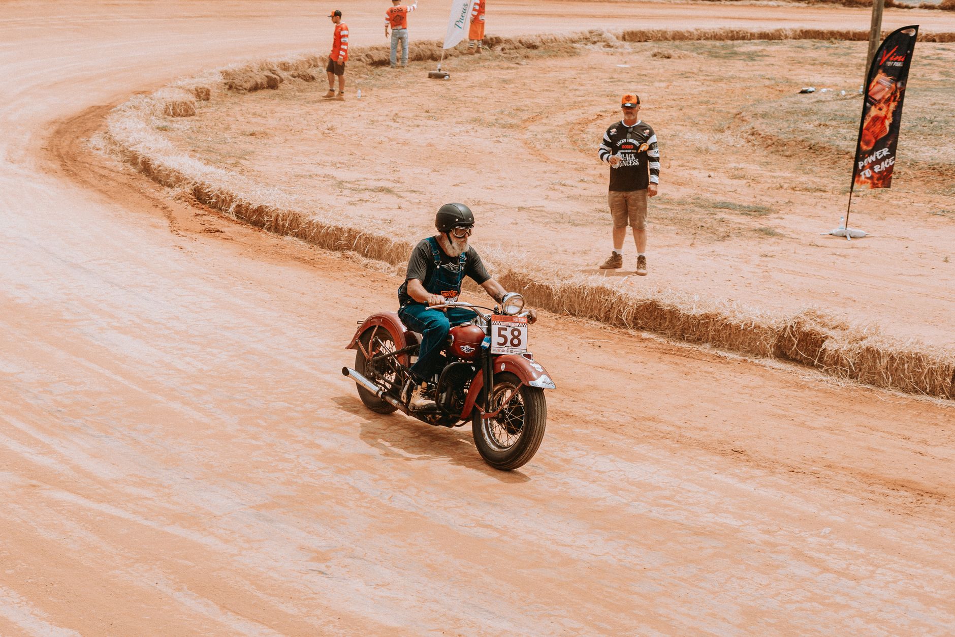 A classic motorcycle rider speeds through a dirt racetrack during a race event on a sunny day.