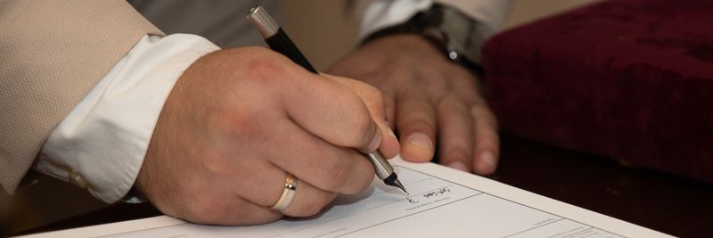 A close-up of a man signing a document, showcasing a wedding ring and pen.