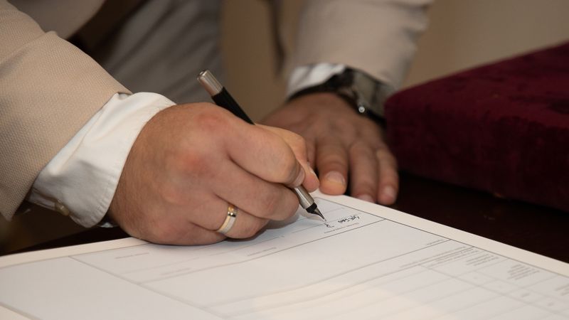 A close-up of a man signing a document, showcasing a wedding ring and pen.