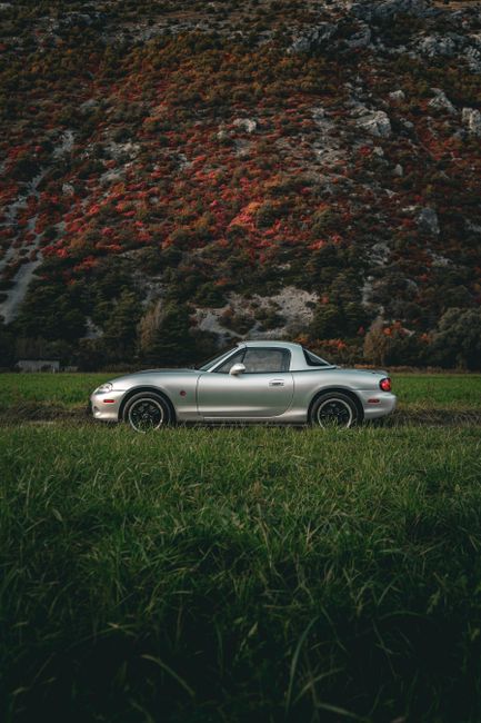 Silver convertible car parked in a lush green field against a mountain backdrop during fall.