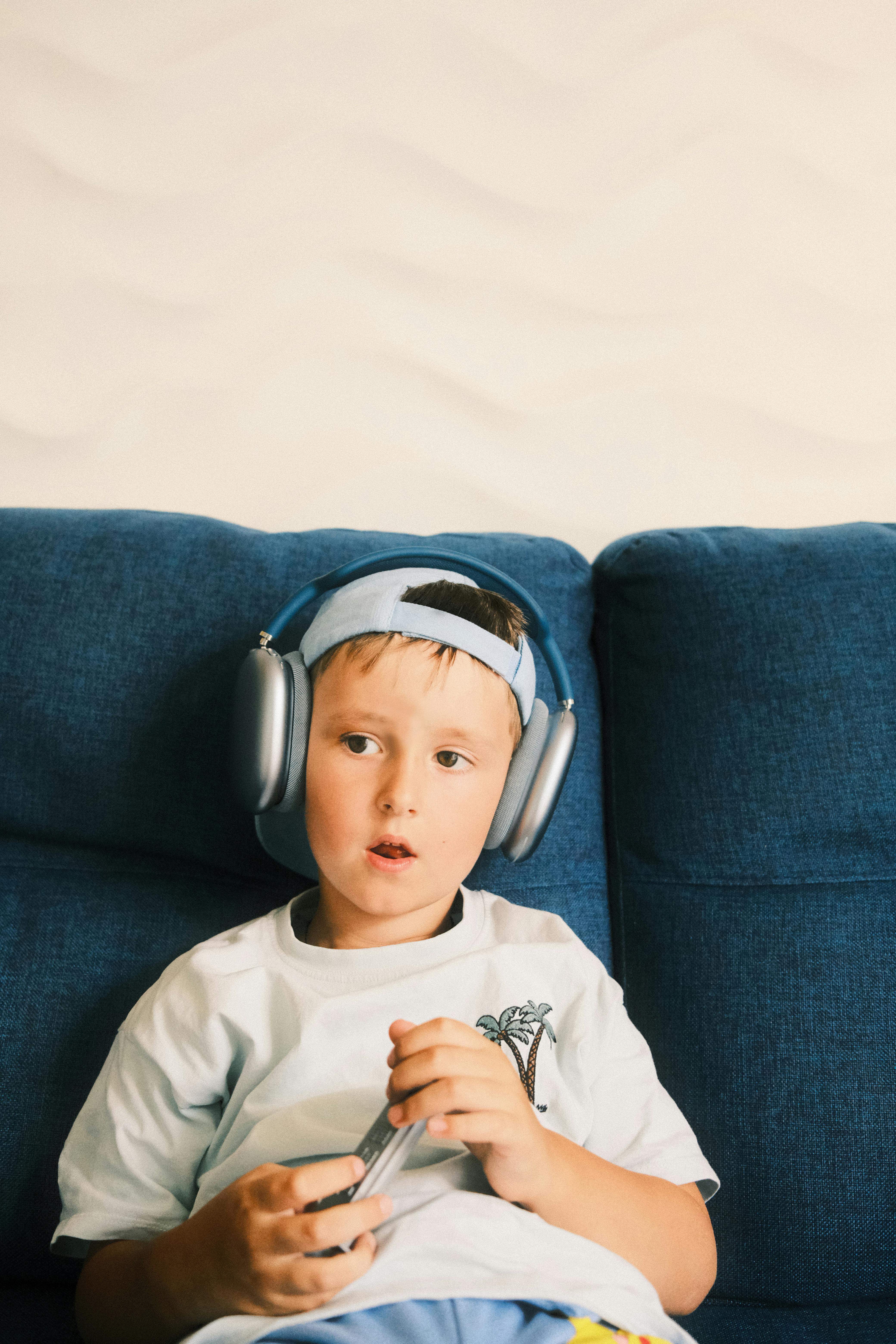 A young boy enjoying leisure time on a sofa, wearing headphones and holding a phone.