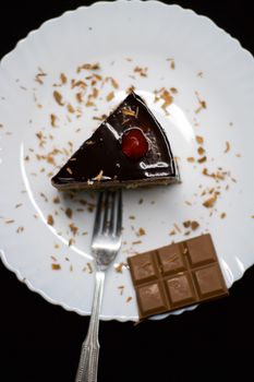 A piece of rich chocolate cake topped with a cherry on a plate with chocolate bar.
