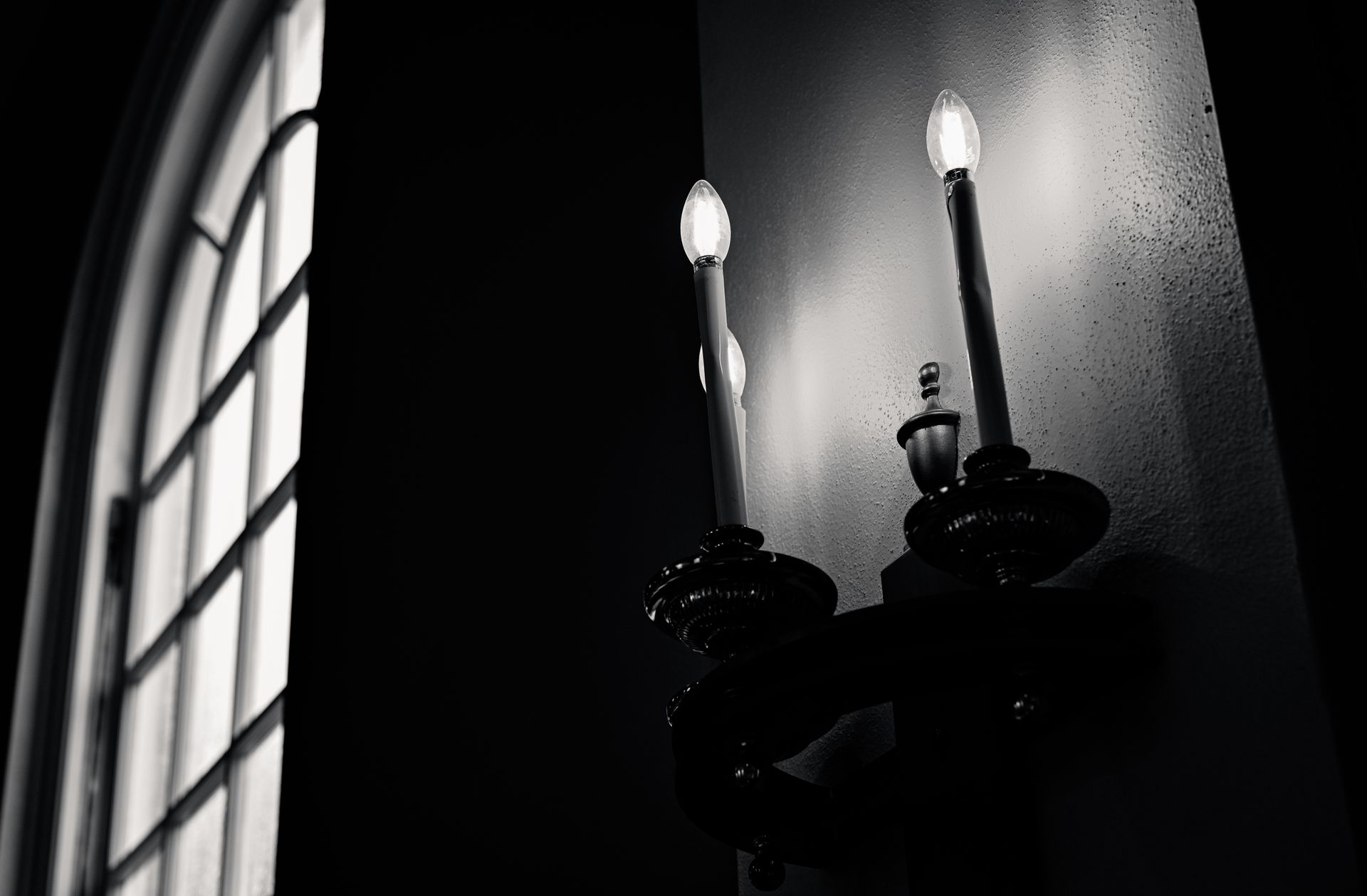 Black and white image of a vintage wall sconce with candles near an arched window.
