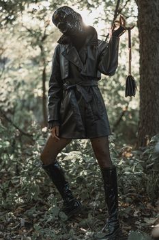 Woman in leather mask and outfit stands mysteriously in a forest, bathed in sunlight.