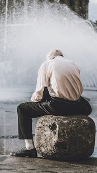 一位老人坐在喷泉旁的巨石上冥想，身影平静地倒映在户外