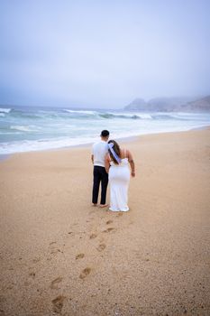A couple walks arm-in-arm along a serene beach, leaving footprints in the sand.
