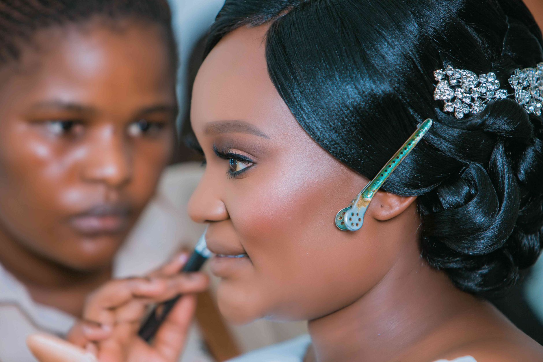Close-up of a woman with intricate hairdo and makeup, assisted by a makeup artist indoors.