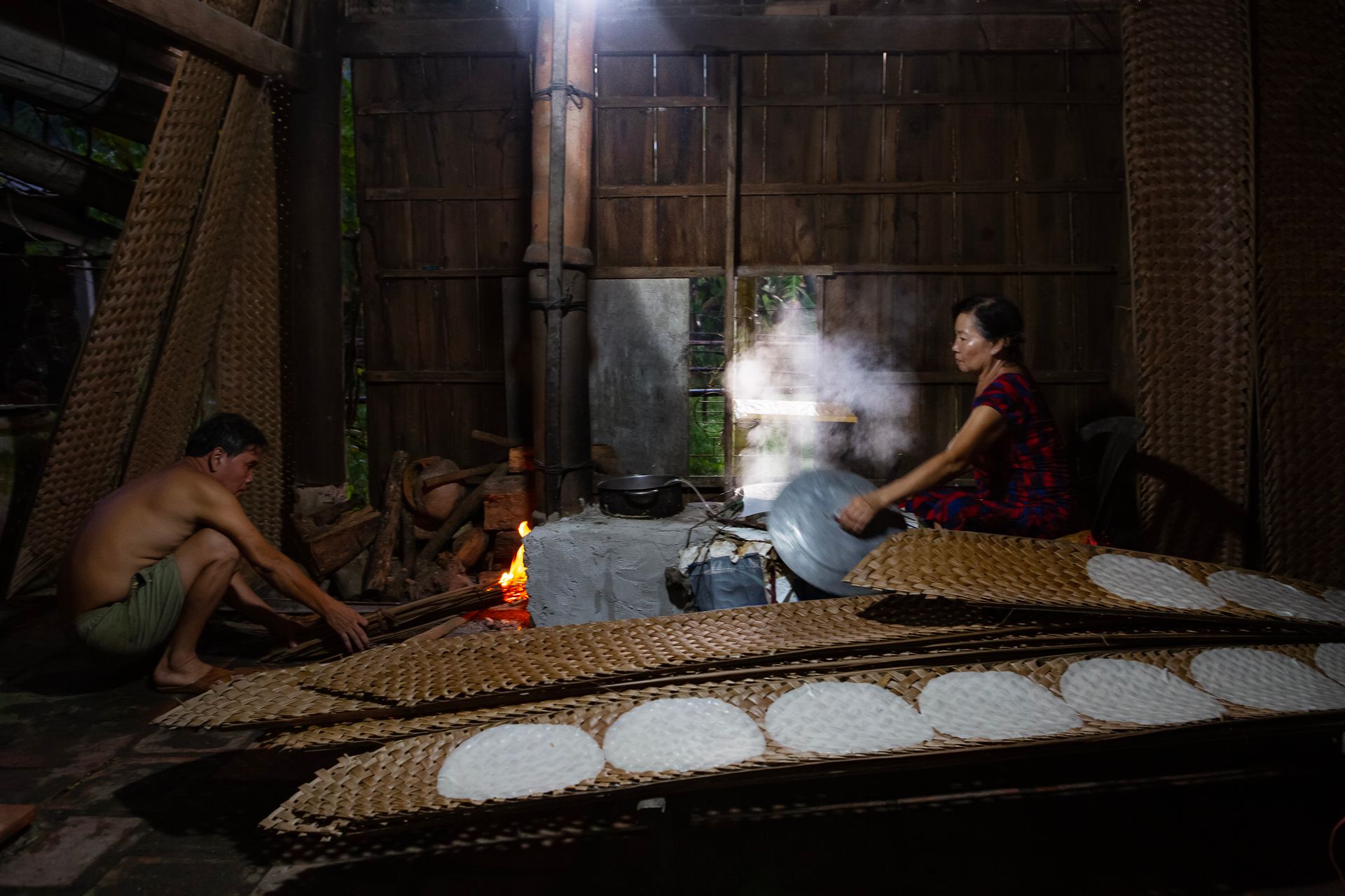 A man and woman making Vietnamese rice paper with a stove inside a rustic hut.