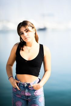 A young woman in trendy fashion poses confidently by a serene seaside backdrop.