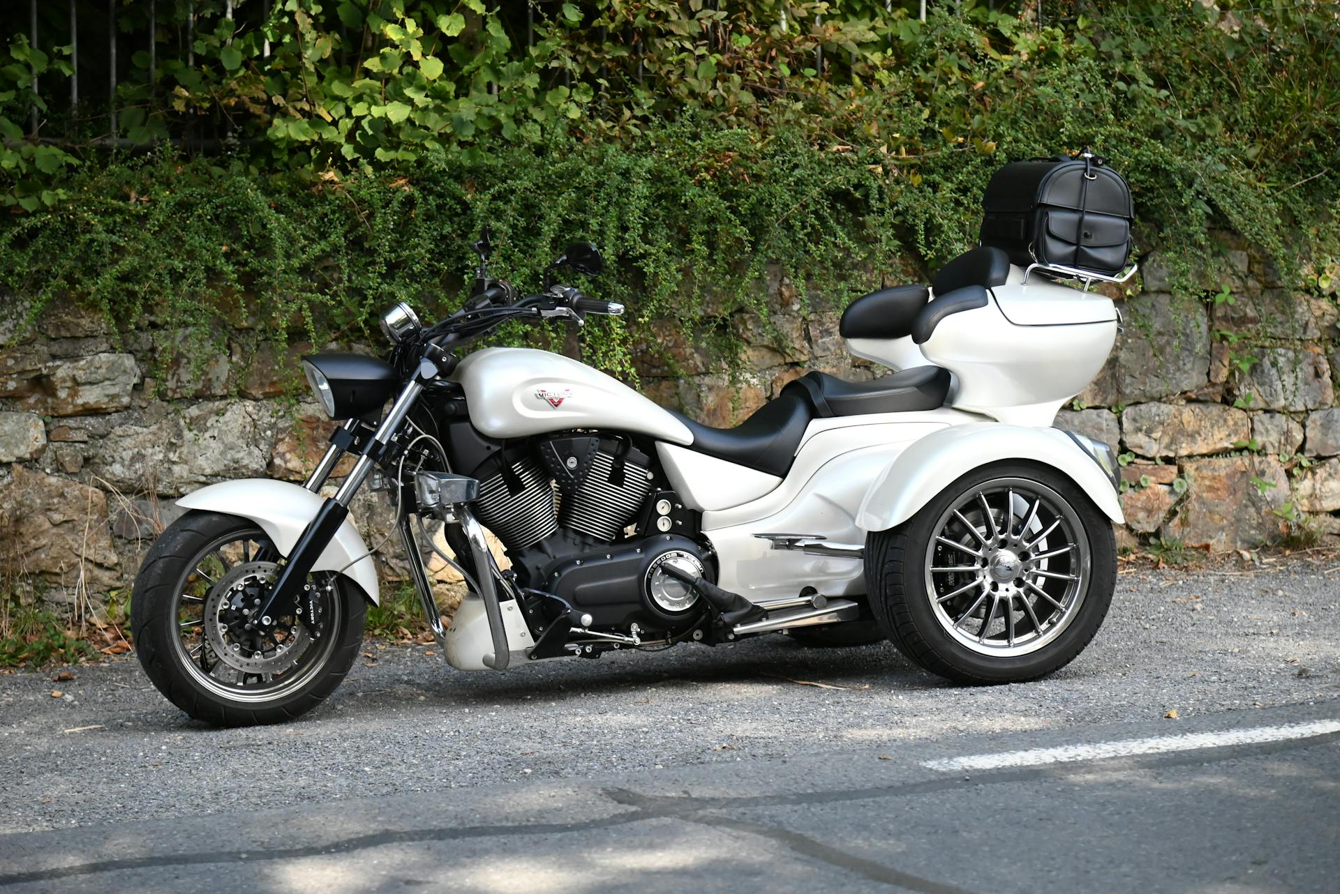 A sleek white trike motorcycle parked against a stone wall with lush greenery background.