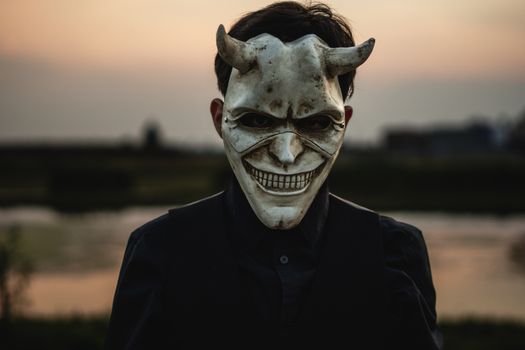 A person wearing a devil mask with horns against a sunset backdrop, creating a spooky atmosphere.