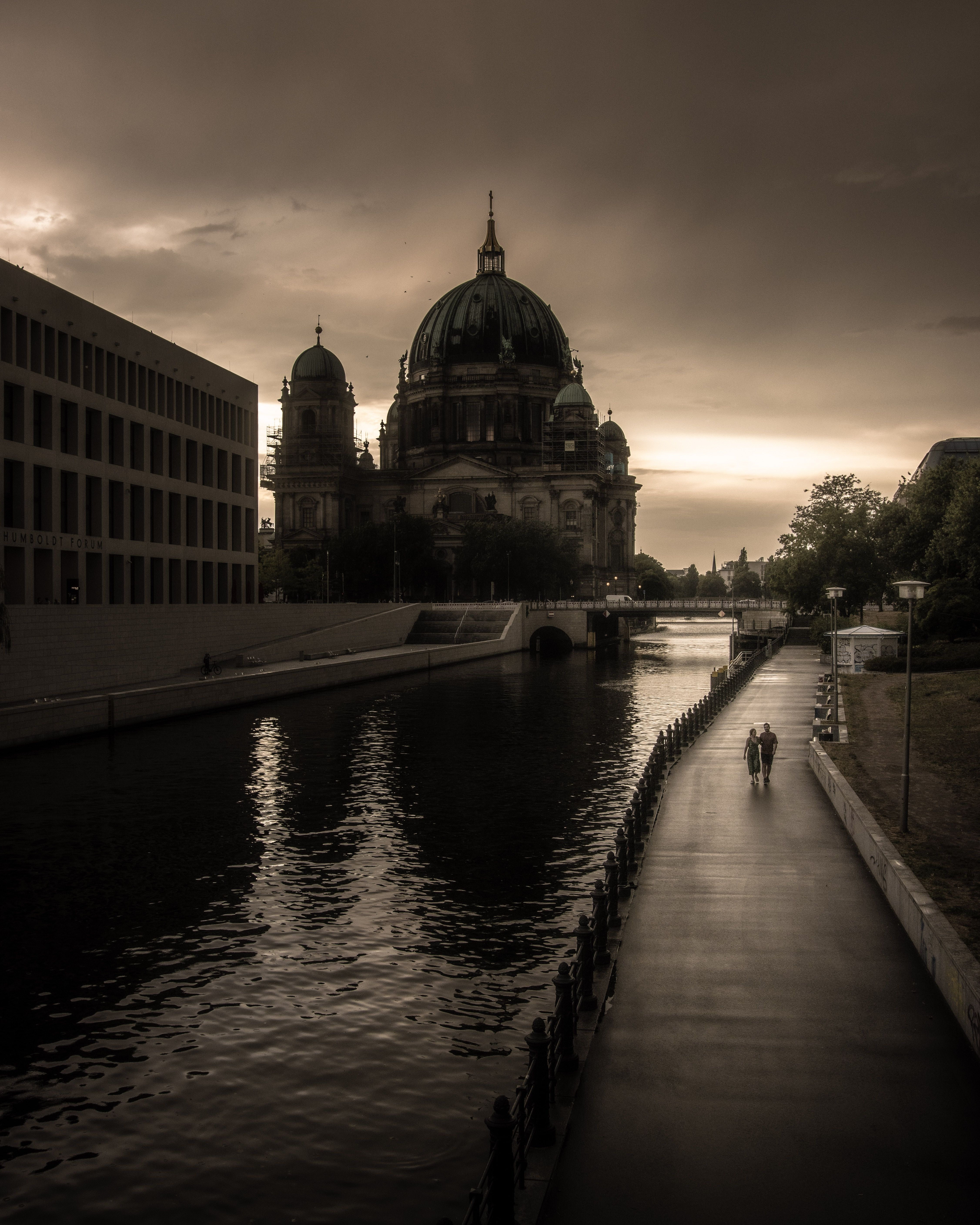 Couple walking by Berlin Cathedral along riverbank in moody dusk lighting.