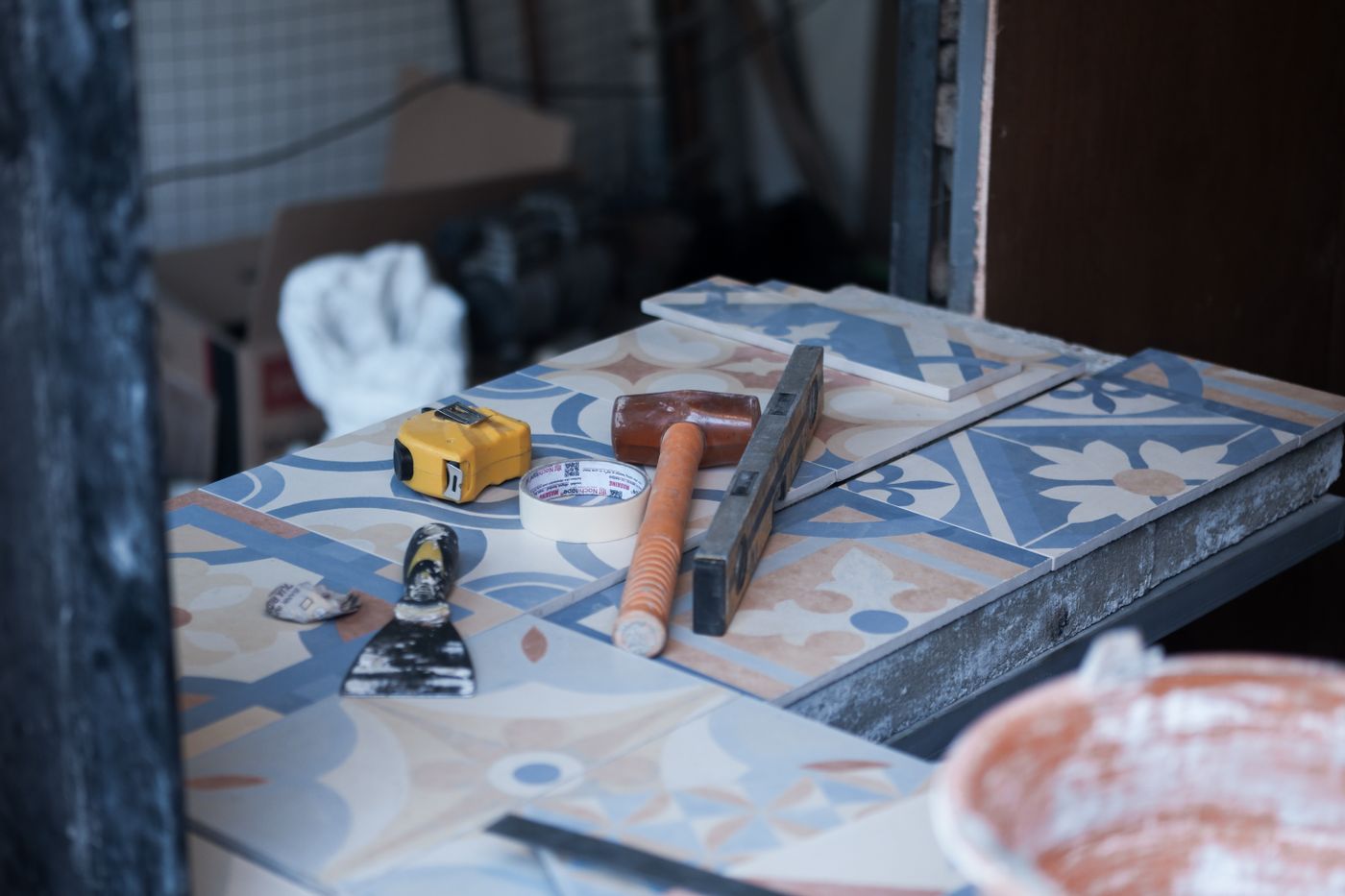 Tiling tools arranged on patterned tiles, including a tape measure, mallet, straightedge, scraper and masking tape.