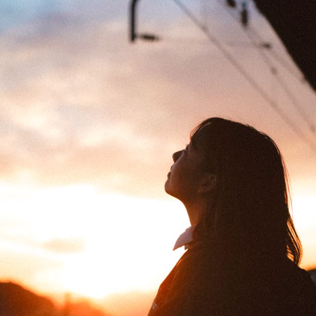 A silhouette of a girl gazing upwards against a vibrant sunset sky, under industrial structures.