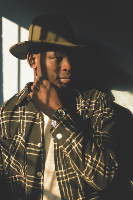 Handsome man wearing a fedora and plaid overshirt, illuminated by dramatic shadows.