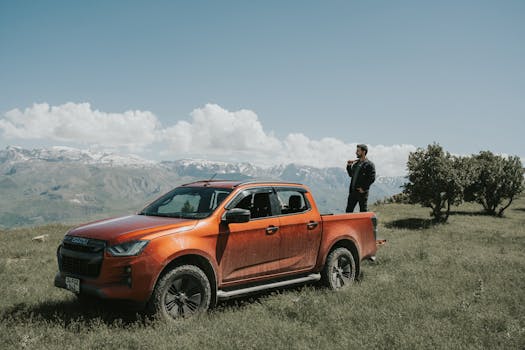 Man with orange pickup truck enjoys picturesque mountain view under a sunny blue sky.