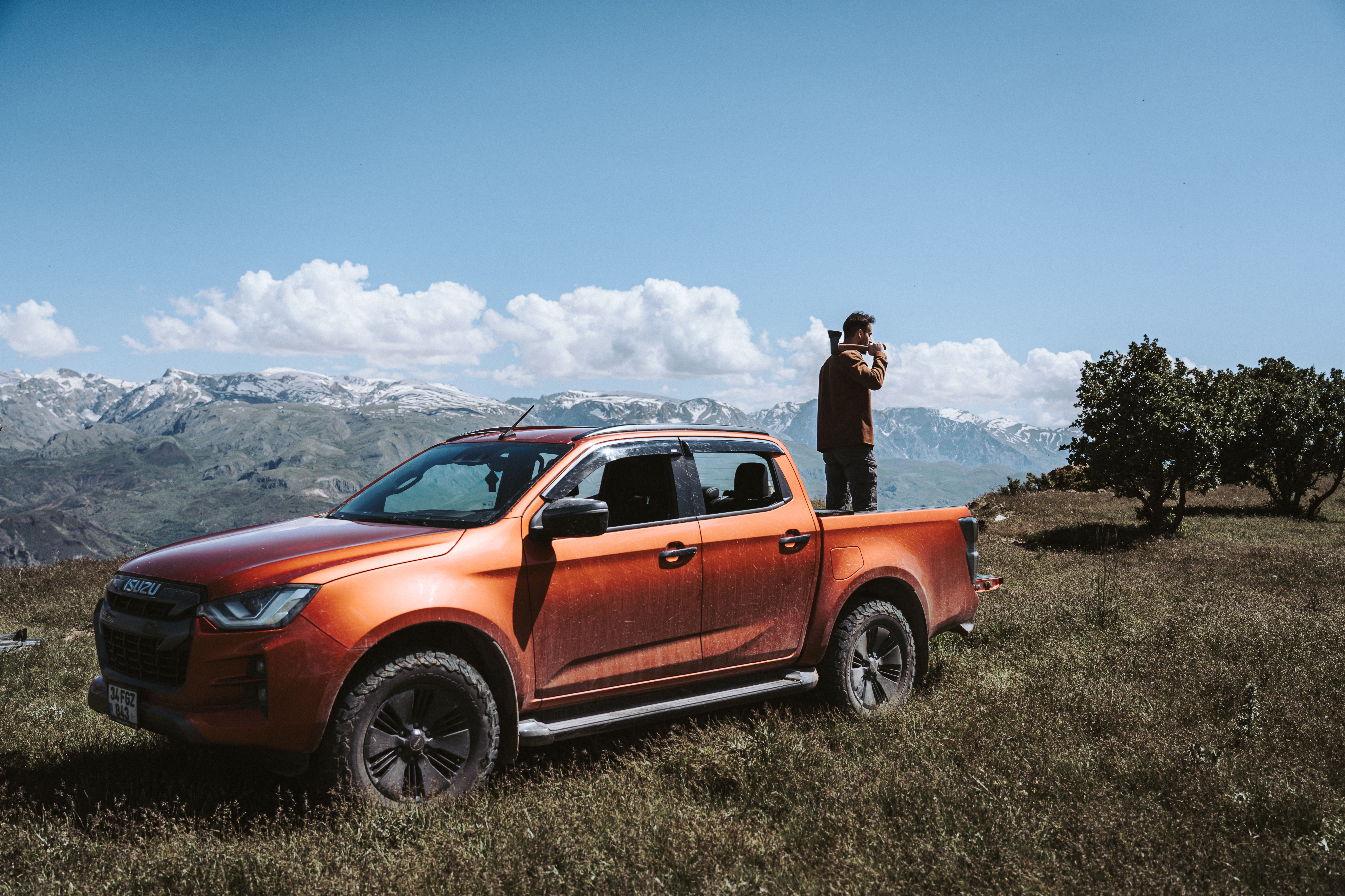 Man stands on orange pickup truck in a picturesque mountain field under a clear blue sky.