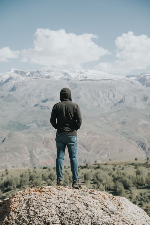 Free A person in a hoodie stands on a rock, overlooking a vast mountainous landscape under a blue sky. Stock Photo