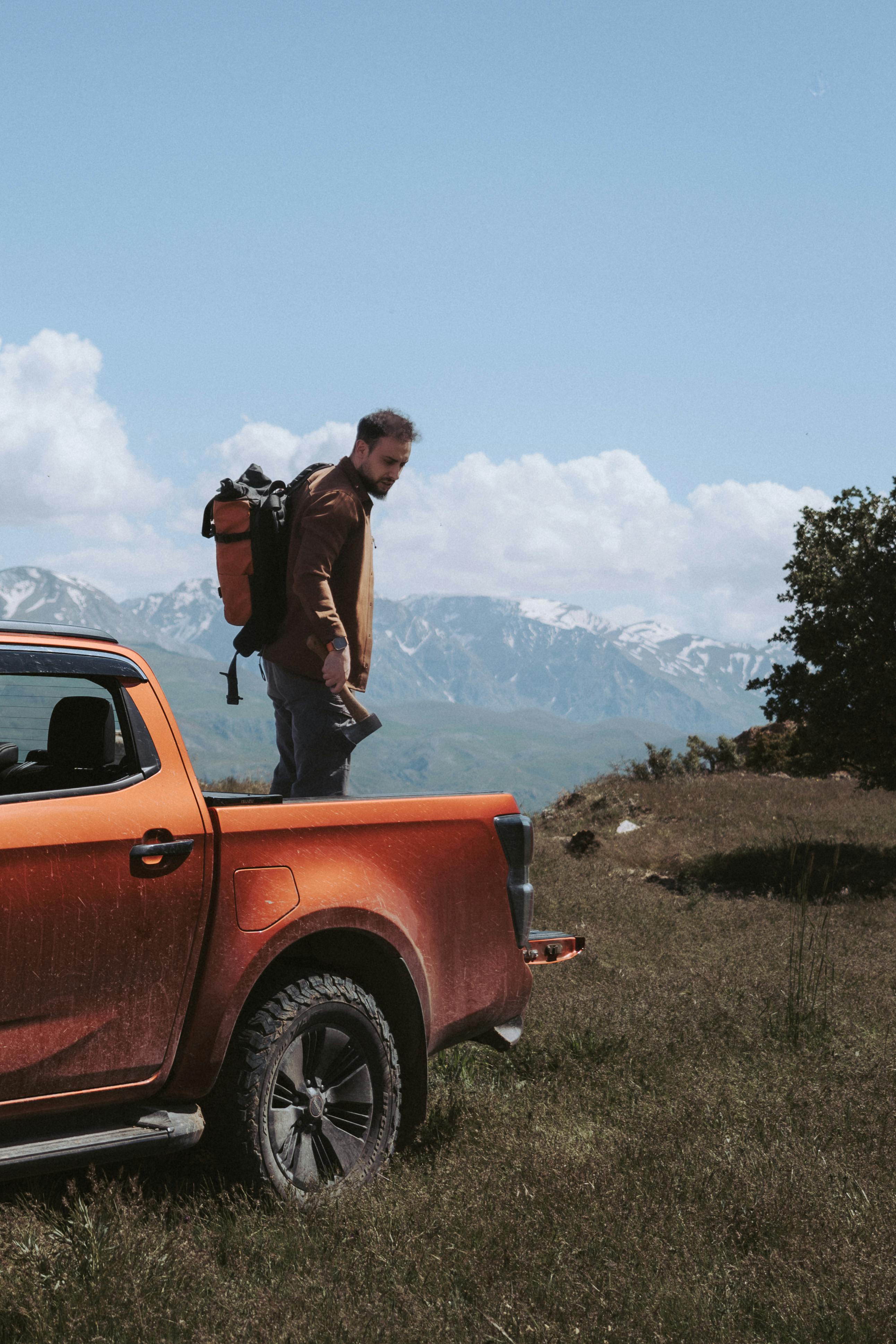 Man with backpack and hatchet exploring mountains beside a pickup truck.