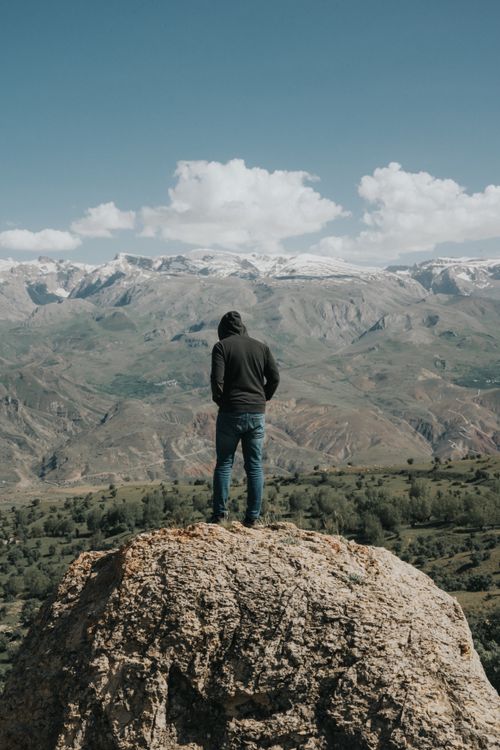 Free A lone man stands on a rocky cliff, gazing at the expansive mountain valley under a clear blue sky. Stock Photo