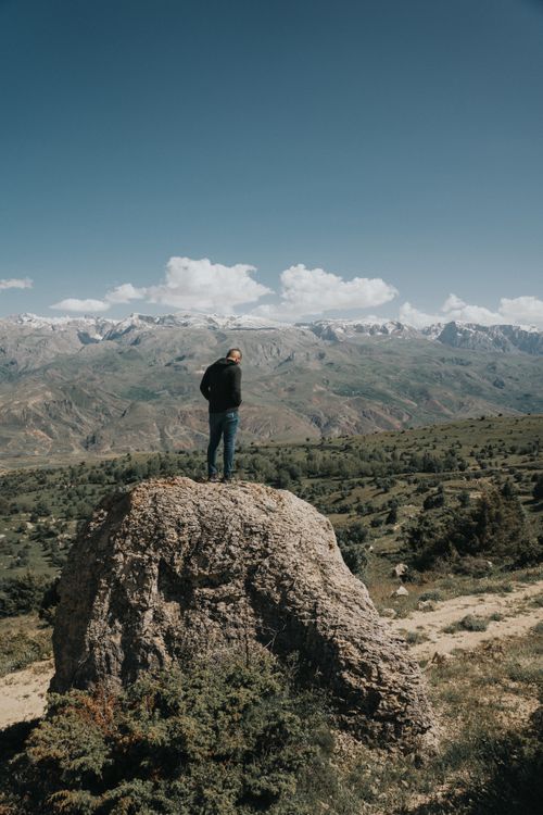 Free A solitary man stands on a rock, gazing at a vast mountain range under a clear blue sky, capturing a moment of reflection. Stock Photo