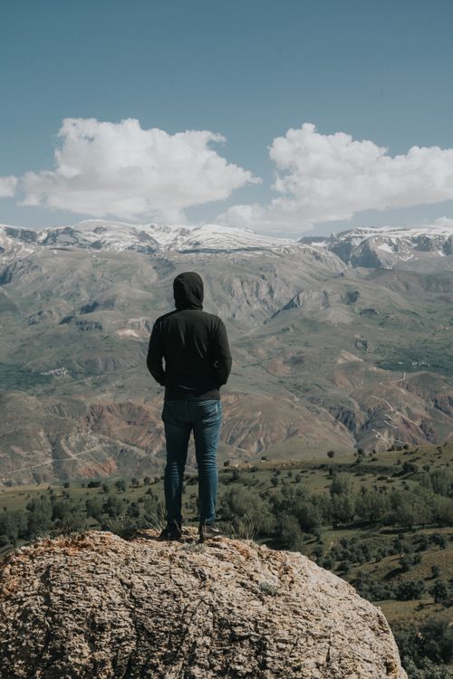 Free A person stands on a rock, observing a stunning mountain valley under a blue sky, suggesting exploration and freedom. Stock Photo
