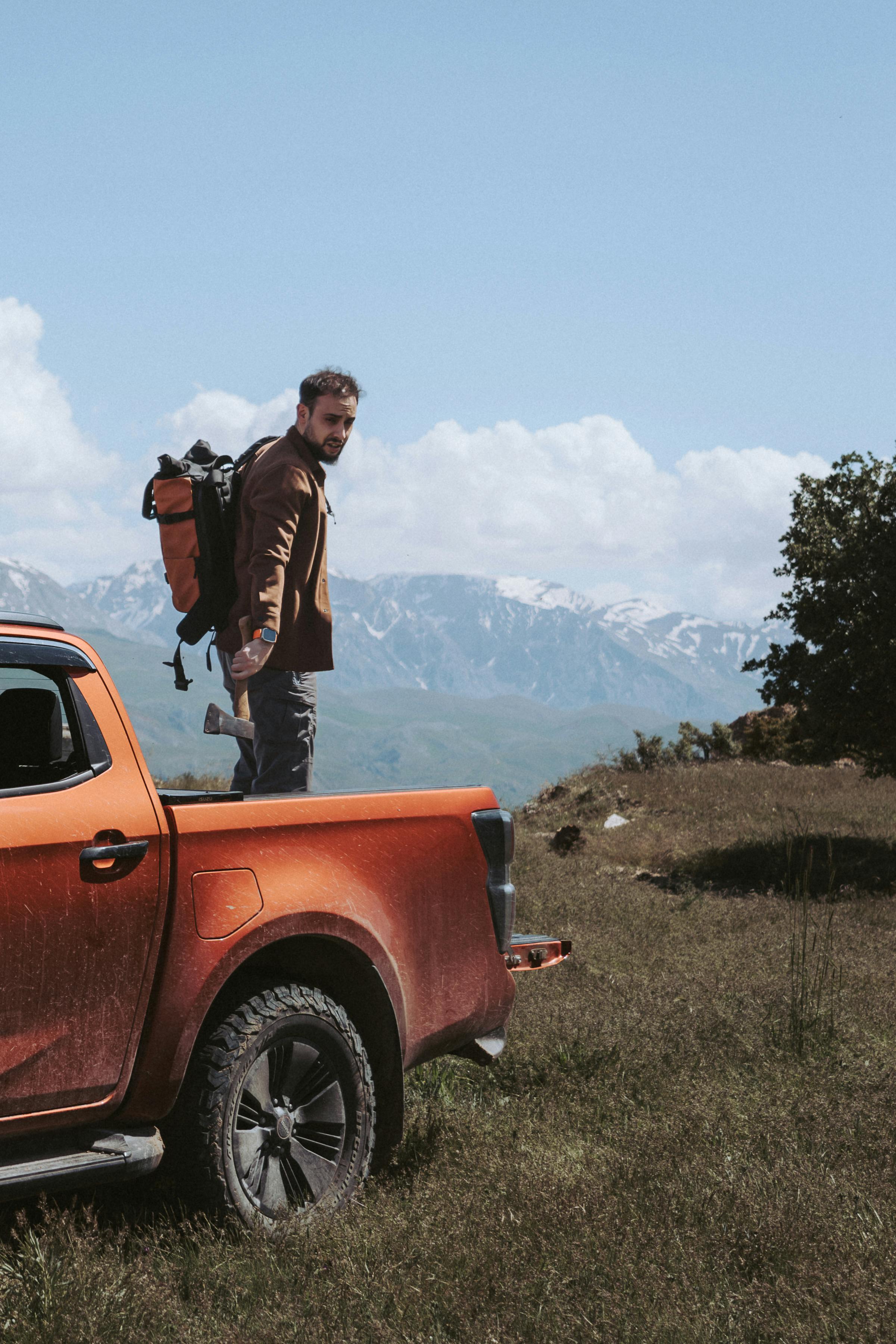 Man with backpack standing by pickup truck, ready for outdoor adventure in mountainous landscape.