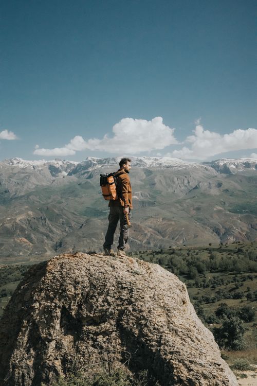 Free Man standing on a rock with a backpack, overlooking scenic mountain landscape on a clear day. Stock Photo