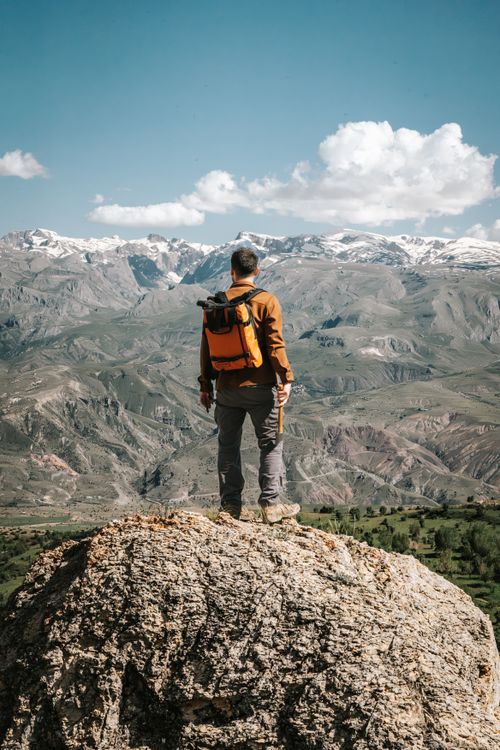 Free A man with a backpack stands on a rock with a scenic view of snow-capped mountains under a clear blue sky. Stock Photo