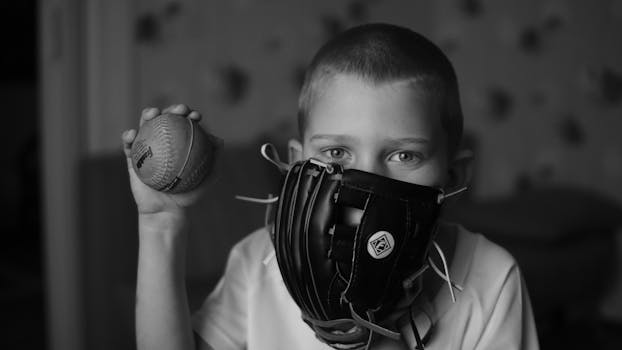 A black and white portrait of a young boy holding a baseball and wearing a glove indoors.