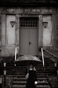 A person stands on steps holding an umbrella, evoking a mysterious, gothic atmosphere.
