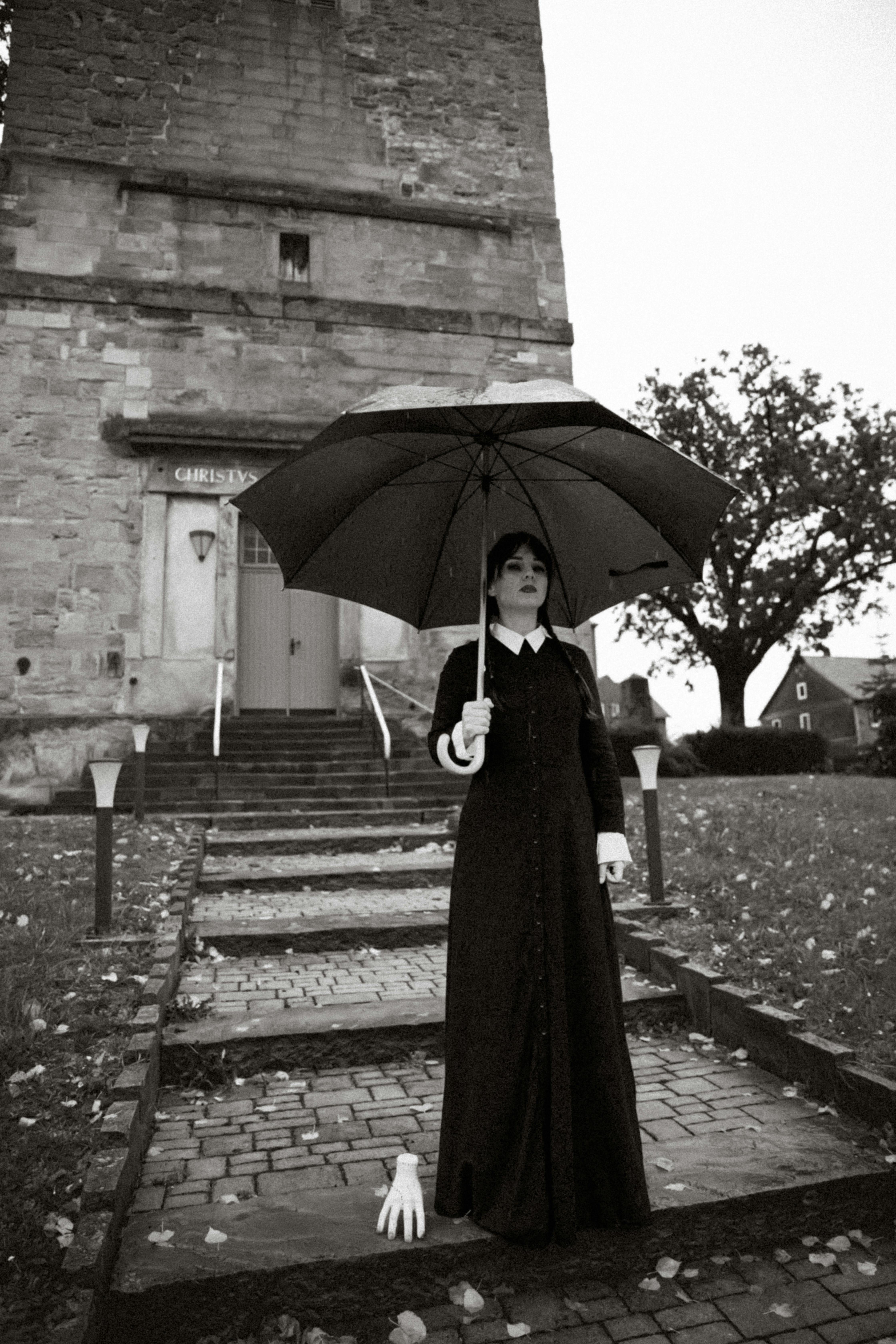 A spooky gothic woman holding an umbrella stands on a cobblestone path in front of an old building.