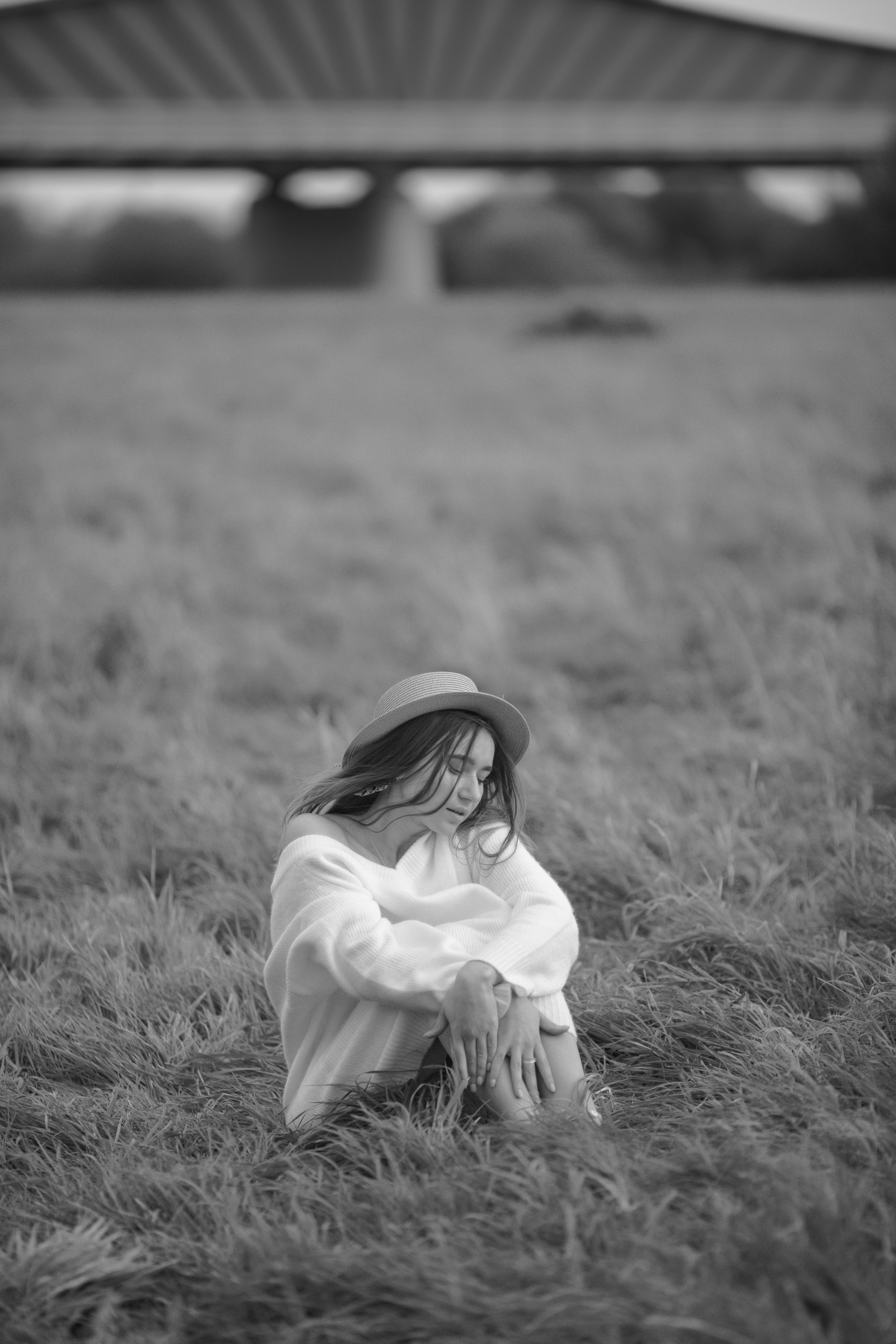 A woman in a hat sitting calmly in a field with a bridge in the background, black and white image.