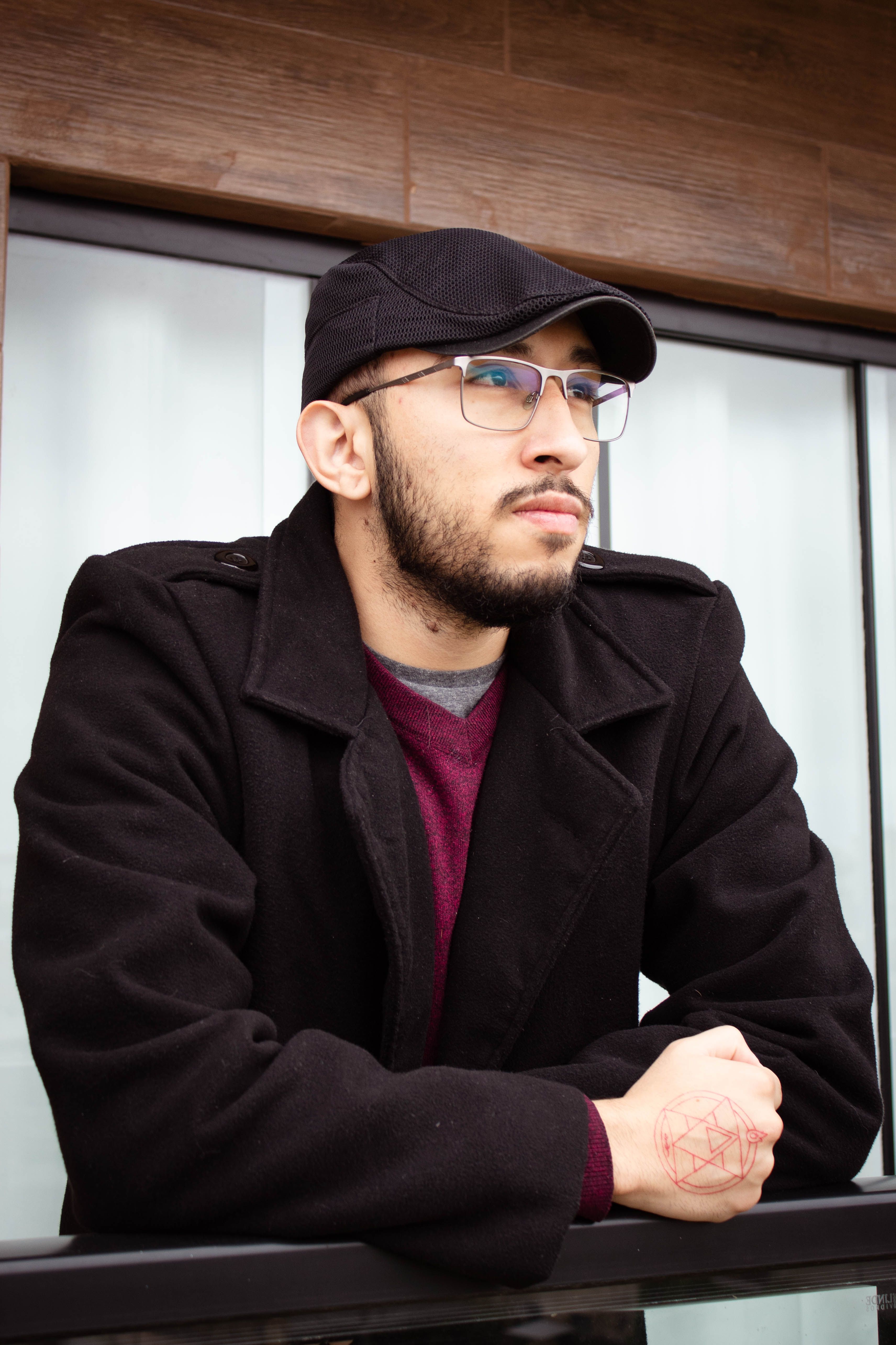 A pensive man with a beard and eyeglasses standing on a balcony, wearing a black coat and flat cap.
