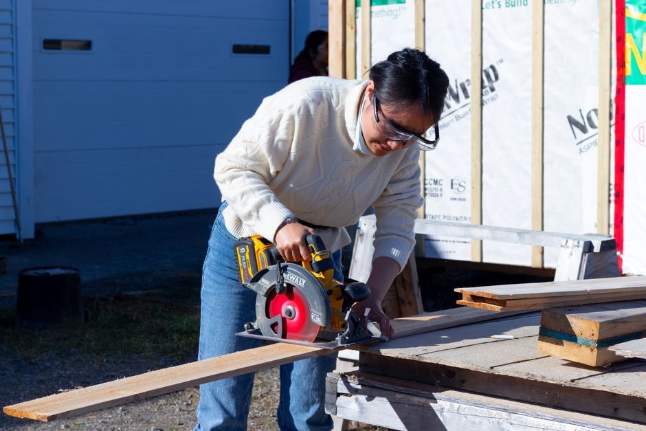 A woman carefully uses a circular saw to cut wood outdoors during a construction project.