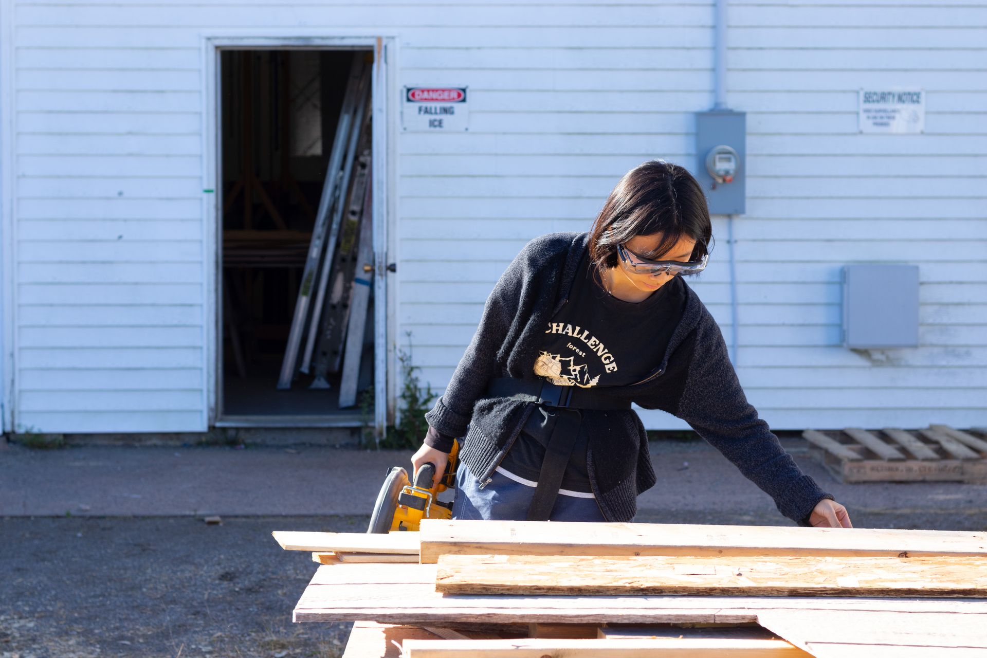 Female carpenter working with a cordless circular saw on wood outdoors, wearing protective glasses.