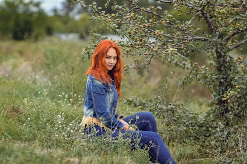 Red-haired Model in a Denim Jacket and Jeans Sitting in a Meadow