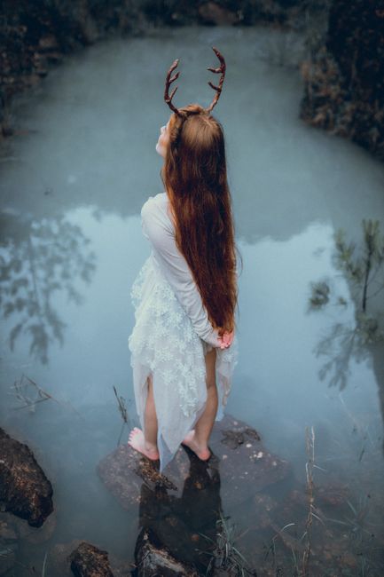 A red-haired woman with antlers poses barefoot on rocks near a mystical river, wearing a white lace dress.