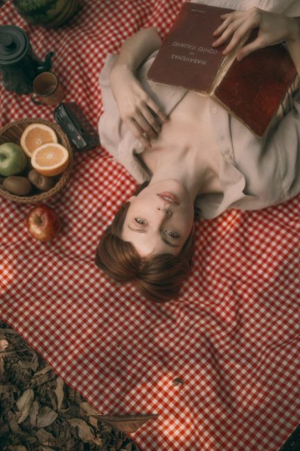 A young woman lies on a picnic blanket, surrounded by fruits and reading a book outdoors.