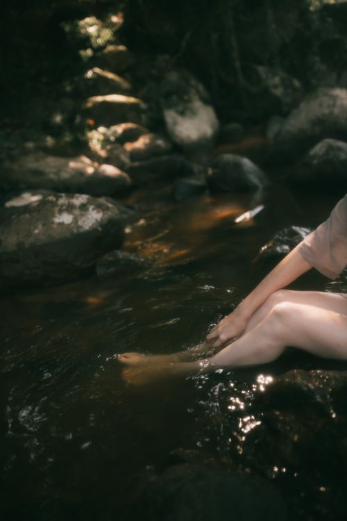 Free A woman sitting by a tranquil stream, dipping her feet in the water surrounded by stones. Stock Photo