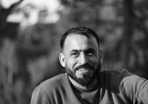 Free A candid black and white portrait of a smiling man with a beard, set against a blurred outdoor background. Stock Photo