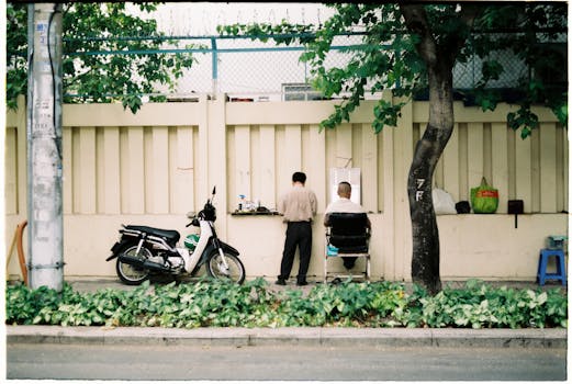 Two men at a barbershop setup on a city sidewalk with a motor scooter nearby.