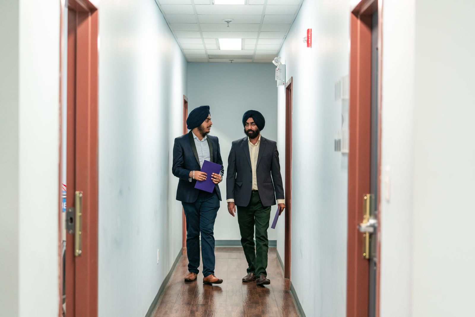 Two men in suits and turbans walking and talking in an office corridor.