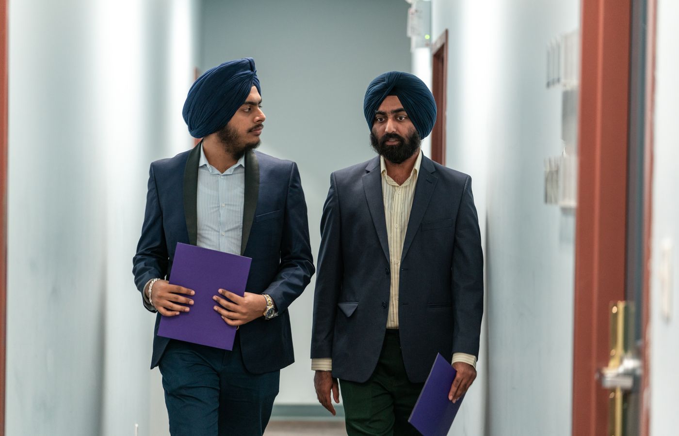 Two businessmen in turbans discuss work while walking through an office hallway.