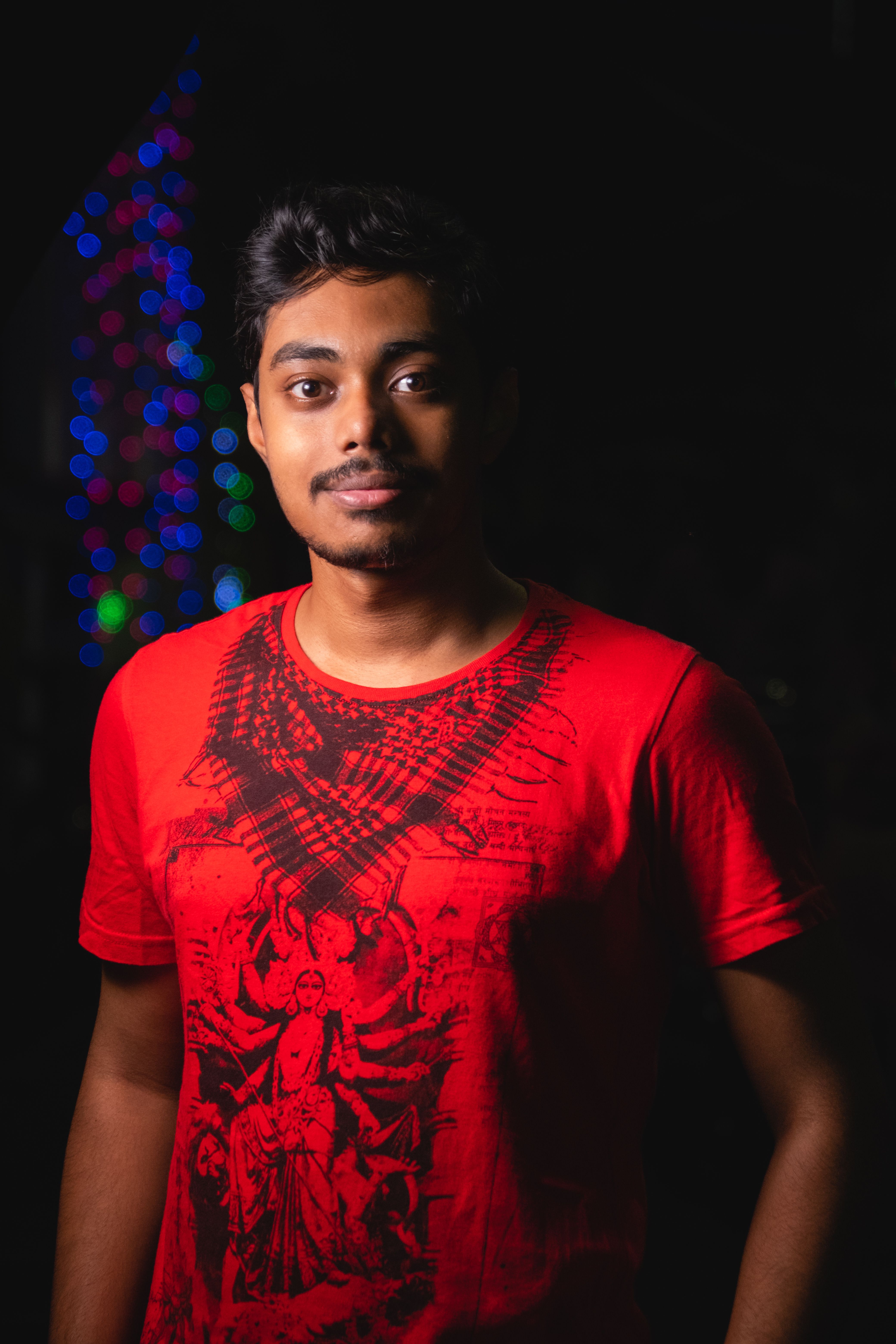 A cheerful man in a red t-shirt standing against a colorful bokeh background at night.